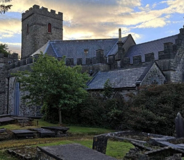 A stone church with a crenellated tower and slate roof is surrounded by greenery and gravestones under a partly cloudy sky.